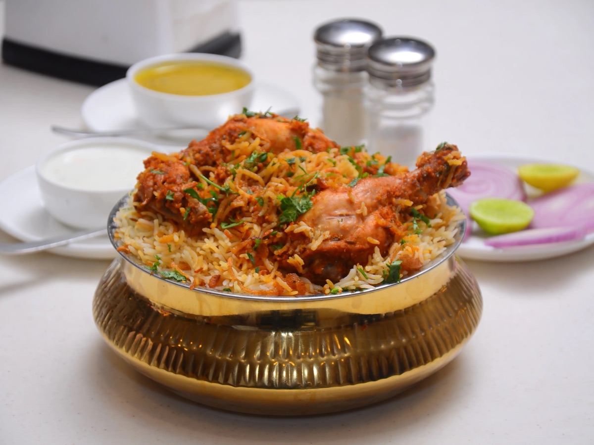 Copper handi of dum biryani just opened at the table, rice and saffron steaming beside a plate of raita and mirchi ka salan